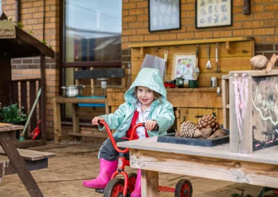 Child riding a tricycle in the outdoor mud-kitchen play area at Little Gems preschool